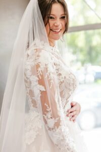 Weddings With Joy Bride wearing long veil and dress smiling at herself in the mirror.