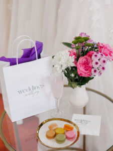 Small glass table with macroons and champagne seated on top. There is also a small gift bag and bouquet of pink flowers.