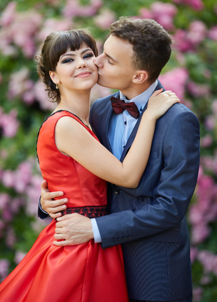 Prom couple dressed to the nines while embracing. The young man is wearing a blue suit with a burgandy bowtie to match his date's prom dress.