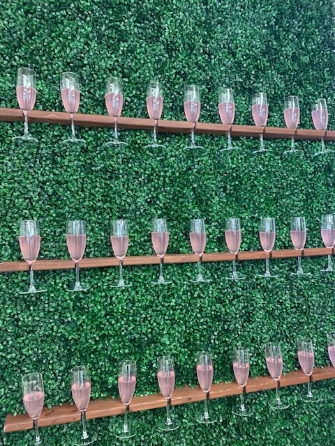 Wedding With Joy's champagne wall with lush greenery for the background and flutes of pink champagne displayed in front.