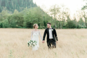 Real Bride and Groom from Weddings with Joy walking hand in hand through a grass field while smiling at each other.