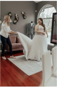 Beautiful, plus-size bride smiling ear-to-ear as her bridal stylist fans out the train on her wedding dress.