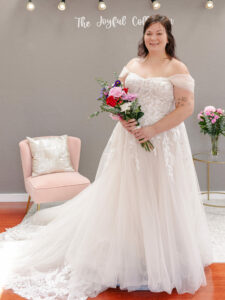 Plus size Weddings With Joy bride standing in her off the shoulder, lace bodice wedding gown while holding a bouquet of flowers.