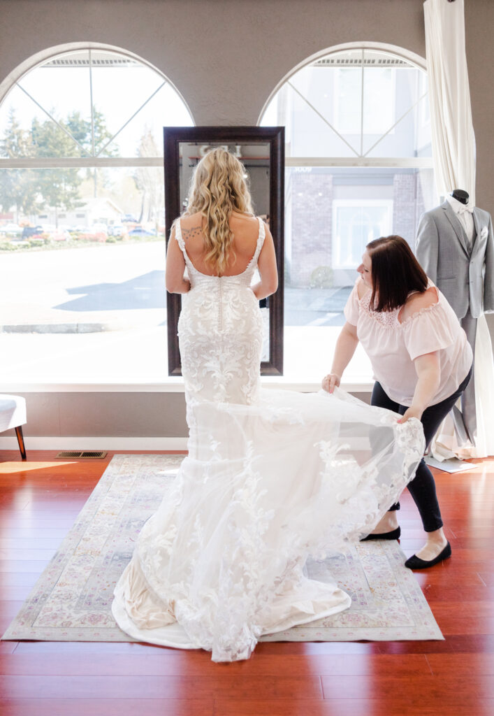 Bridal stylists adjusts wedding dress train while a bride is trying on a wedding gown during shopping appointment. Weddings With Joy. 