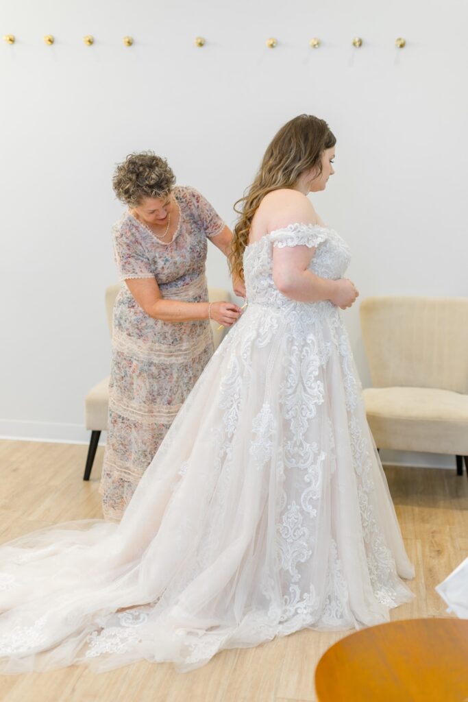 Mother of the bride helps bride button up lace ball gown wedding dress with off shoulders straps and long train. Weddings with Joy. 