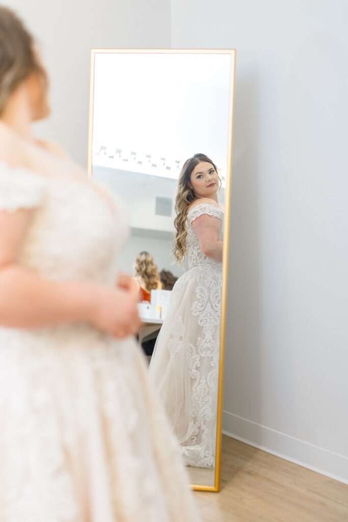 Plus size bride in lace ball gown wedding dress with off the shoulders straps looks in mirror on wedding morning. Weddings with Joy.