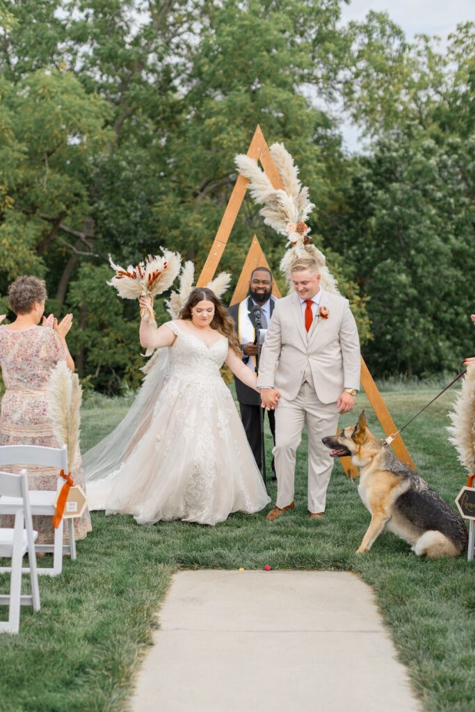 Boho outdoor wedding ceremony with wooden arch backdrop, bride in lace ballgown with off shoulders straps and dried floral bouquet. Weddings with Joy. 