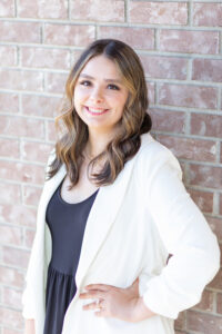 Bridal Stylist from Weddings with Joy, Hannah. She has medium brown hair with blonde highlights and is wearing a black outfit with a white sport jacket.