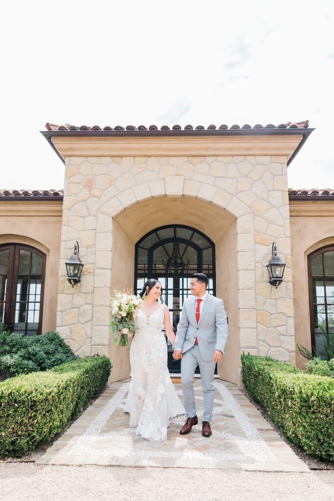 bride in lace wedding dress with straps, long train walks with groom in light grey suit. Weddings with Joy.