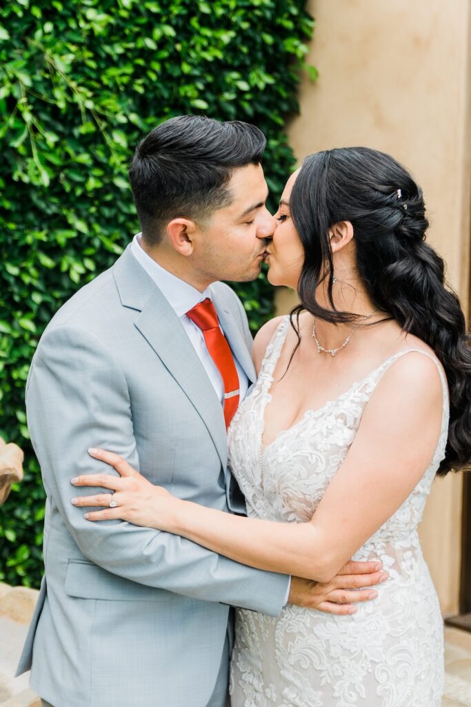 bride in classic lace wedding dress with v-neck and straps kisses groom in light grey suit and burnt orange tie. Weddings with Joy. 