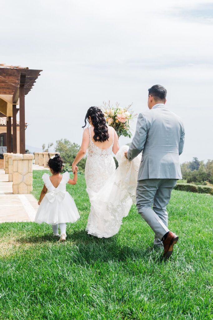 Groom in light grey suit carries bride's dress train. Open back lace wedding dress. Flower girl in white dress with bow on back. Weddings with Joy. 