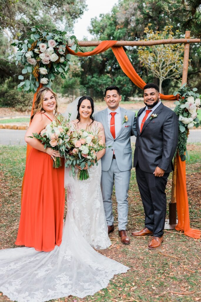 bride in classic lace wedding dress with long dress train. Bridesmaid in burnt orange dress. Floral wedding ceremony arbor. Weddings with Joy.