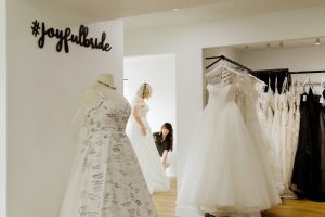 bridal stylist helps woman try on wedding dress and surrounded by dresses on racks